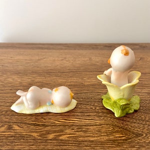 May include: Two porcelain figurines of babies. One baby is lying on a yellow leaf, the other is sitting inside a yellow flower with green leaves.