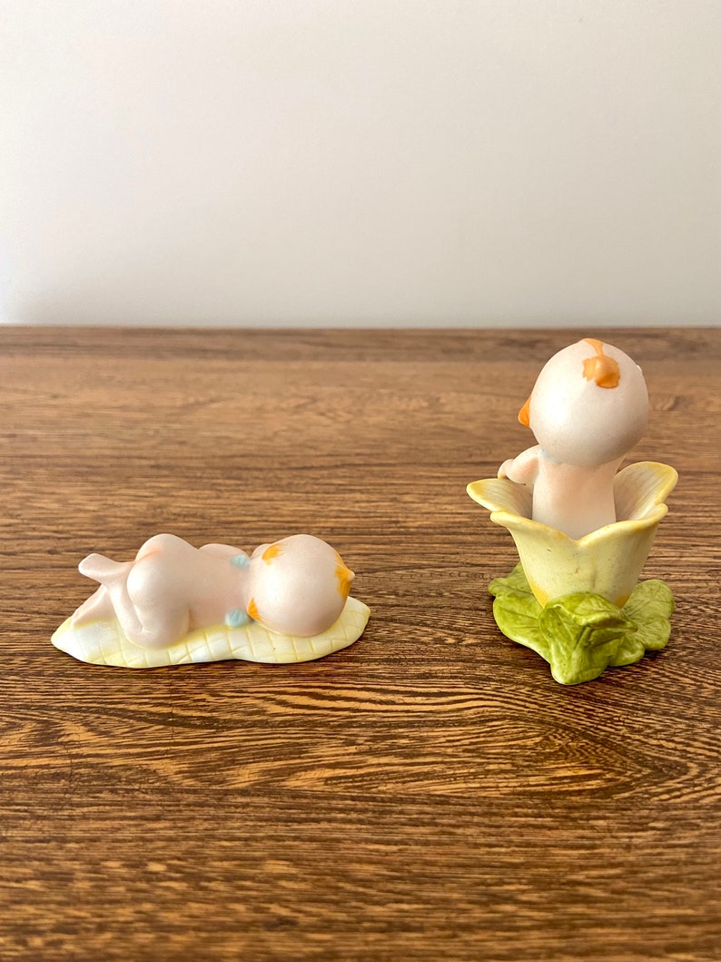 May include: Two porcelain figurines of babies. One baby is lying on a yellow leaf, the other is sitting inside a yellow flower with green leaves.
