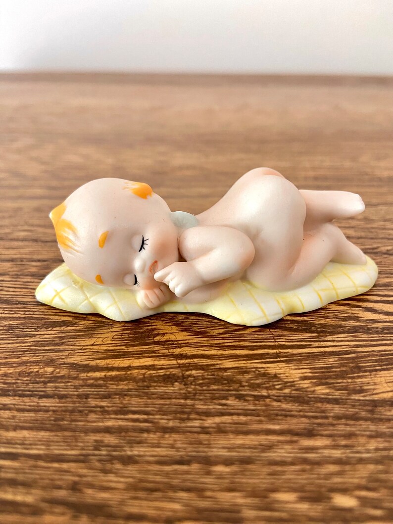 May include: A porcelain figurine of a sleeping baby with yellow hair lying on a yellow and white patterned blanket.