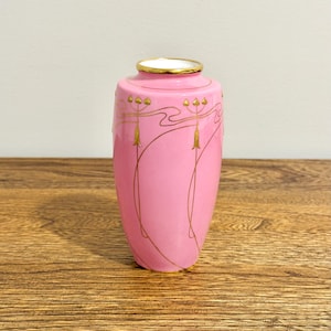 May include: A cylindrical pink vase featuring a gold-coloured rim and delicate gold-coloured line work depicting stylised floral motifs that wrap around the upper portion of the body. The vase has a smooth, glossy finish and a white interior.