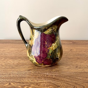 May include: A ceramic pitcher featuring a dark green and black background with a hand-painted design of purple grapes and green leaves. The interior of the pitcher is white, and it has a curved handle and a pouring spout on the upper rim.