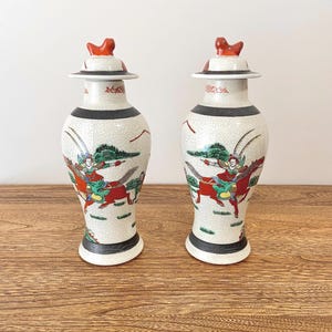 Peut inclure: Deux vases en céramique assortis avec couvercles. Les vases blanc cassé ont un glaçage craquelé et une scène peinte d'une personne à cheval. Les couvercles ont un fleuron rouge et un bord blanc. Vases décoratifs.