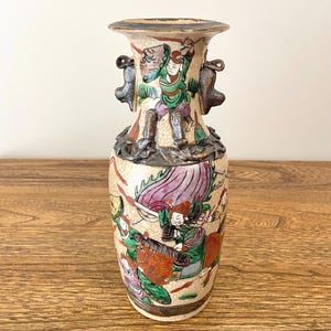 Puede incluir: Jarrón de cerámica vintage con un fondo beige moteado y figuras pintadas a mano en verde, morado y rojo. El jarrón tiene dos asas ornamentadas y un borde acampanado. La obra de arte representa una escena de batalla.