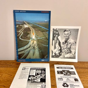 Könnte beinhalten: Ein Schwarzweißfoto eines Astronauten in einem Raumanzug mit einer amerikanischen Flagge im Hintergrund. Der Astronaut lächelt und hält ein Modell eines Space Shuttles. Das Foto ist vom Astronauten signiert, "Col. Mike Mullane 9/21/96".