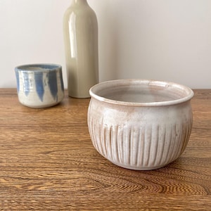 Può includere: Una ciotola in ceramica bianca con un design testurizzato e a coste. La ciotola è appoggiata su un tavolo di legno.