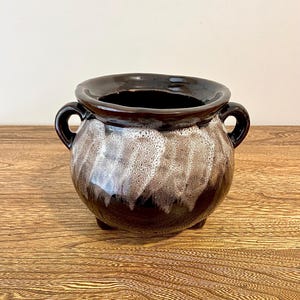 May include: A small brown and white ceramic cauldron with two handles. The cauldron has a speckled glaze and is sitting on a wooden surface.