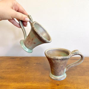 May include: Two handmade ceramic mugs with a unique design. The mugs have a flared shape, a light green base, and a brown and green mottled finish. One mug is held by a hand, while the other sits on a wooden surface. The mugs have a distinctive handle.
