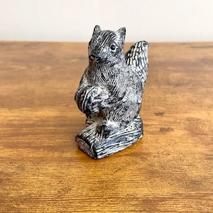 Puede incluir: Una figurita de ardilla tallada en esteatita en blanco y negro que sostiene una nuez, sentada sobre una base de tronco.