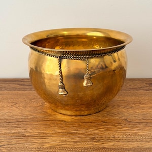 May include: A hammered brass planter with a wide rim and a rounded body. The planter is adorned with a rope detail and tassels. The planter is sitting on a wooden surface.