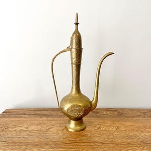 Puede incluir: Una tetera de latón de color dorado con intrincados diseños grabados. La tetera tiene un cuerpo alto y delgado con un pico curvo y un asa.