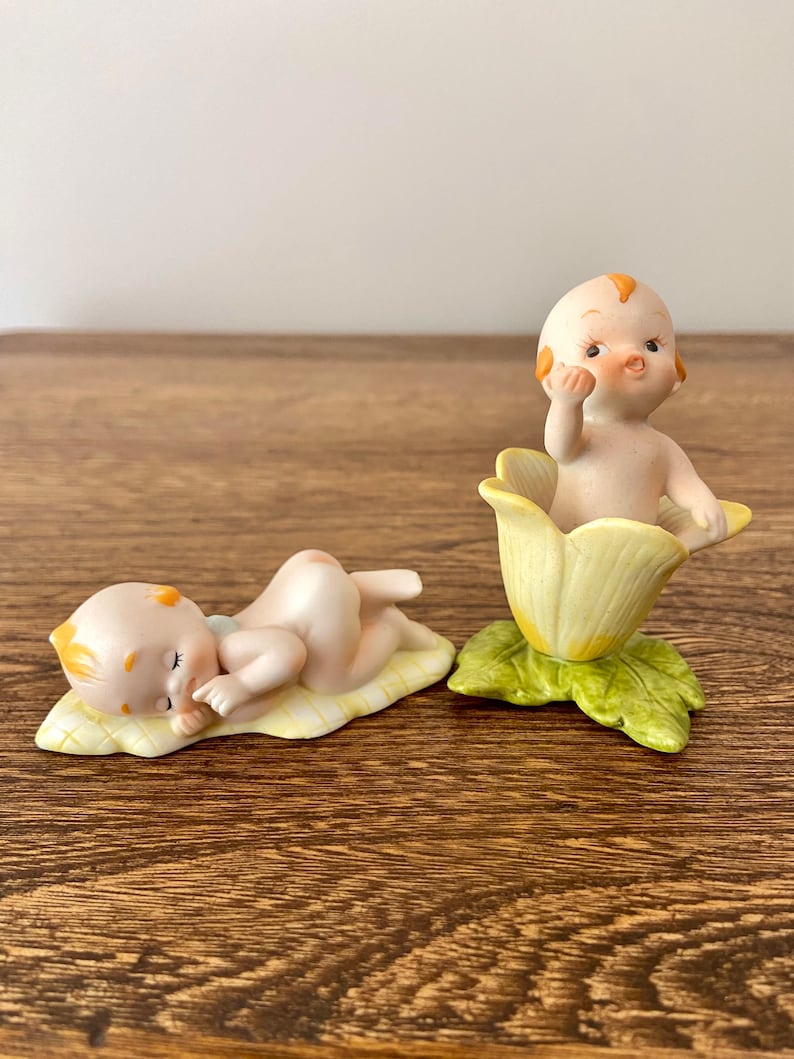 May include: Two porcelain figurines of babies. One is sleeping on a yellow and green leaf. The other is sitting in a yellow flower with green leaves.