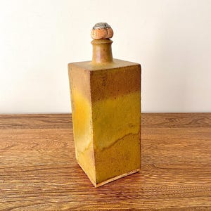 Puede incluir: Una botella de cerámica rectangular hecha a mano con un tapón de corcho. La botella tiene un degradado de tonos amarillos y marrones. El tapón es de color marrón claro con una parte superior texturizada y redondeada. La botella se exhibe sobre una superficie de madera.