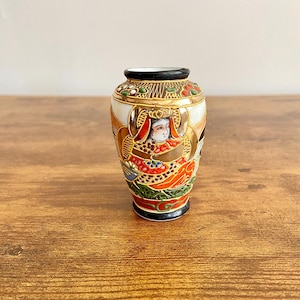 Puede incluir: Un pequeño jarrón de porcelana blanca con borde negro y detalles dorados. El jarrón está decorado con una pintura japonesa colorida de una mujer con una túnica roja y dorada.