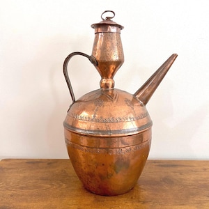 Puede incluir: Una jarra antigua de cobre con un pico largo, un asa y una tapa. La jarra tiene un diseño ornamentado grabado en la superficie. El cobre tiene un tono marrón rojizo cálido, y la jarra está sobre una superficie de madera.