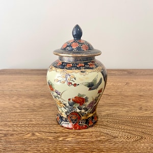 Puede incluir: Un pequeño jarrón de cerámica de color crema con tapa. El jarrón está decorado con un diseño floral y de pájaros en tonos rojos, azules, verdes y dorados. La tapa es negra con detalles dorados.
