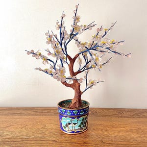 Puede incluir: Un bonsái decorativo con tronco marrón, ramas azules y acentos florales rosas y amarillos. El árbol está plantado en una maceta decorativa azul y dorada con un diseño de grulla. La maceta tiene unos 10 cm de diámetro.