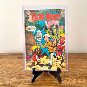 May include: A vintage comic book cover featuring Bob Hope in a jungle setting. The cover art depicts Bob Hope with a group of cartoon characters, including a green monster with an axe, a red-faced monster, and a monkey-like creature. The comic book title is "Bob Hope" and the issue number is "100". The cover text reads "Is there a witch-doctor in the house?"