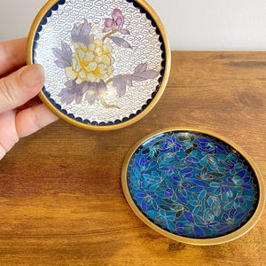 May include: Two decorative coasters with gold rims. One coaster features a floral design with yellow and purple hues on a white background. The other coaster has a blue floral pattern. Both coasters have a gold rim and a dark blue border.