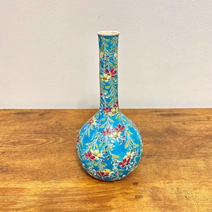 May include: A turquoise ceramic vase with a bulbous base and a long, slender neck. The vase is adorned with a floral pattern in shades of pink, yellow, and green. The vase is displayed on a wooden surface.