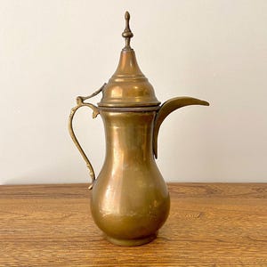 Può includere: Una teiera in ottone con un coperchio e un beccuccio. La teiera ha un design arabo tradizionale.