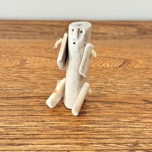 May include: A handcrafted, light-coloured wooden figure with a cylindrical body, rectangular arms, and legs. The figure has two small holes for eyes and is connected with twine. The figure is sitting on a wooden surface.