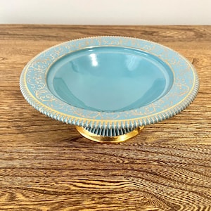 Könnte beinhalten: Eine hellblaue Schale auf einem Sockel mit goldfarbenem Rand und filigranen goldenen Verzierungen. Die Schale steht auf einer Holzoberfläche.