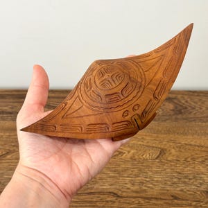 May include: A hand-carved wooden object with a curved, pointed shape and a central circular motif featuring intricate geometric patterns. The wood has a warm, medium-brown tone with visible grain, and the surface is decorated with detailed carvings.