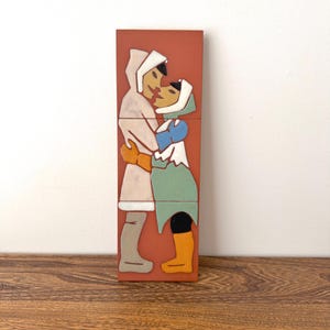 May include: A rectangular, multi-piece tile art piece depicting two figures in winter clothing embracing. The figures are in white, blue, green, and orange, set against a terracotta background. The artwork is on a wooden surface.
