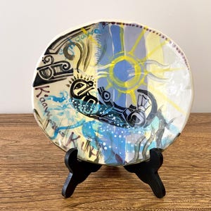 May include: A ceramic plate with a colorful, abstract design featuring a sun, waves, and a stylized creature. The plate is on a black stand and has the words "Kaa dawa" and "Kuna" written on it.