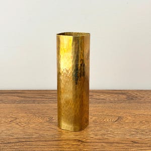 Op de afbeelding: Een hoge, gehamerde messing vaas met een rechthoekige vorm. De vaas heeft een getextureerd oppervlak en een gouden tint, die het licht reflecteert. De vaas staat op een houten oppervlak.