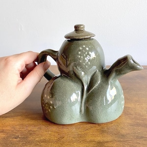 Puede incluir: Tetera de cerámica artesanal en un tono verde apagado. La tetera presenta un cuerpo bulboso con una superficie texturizada y pequeñas motas blancas. Tiene un pico curvo, un asa de bucle y una tapa pequeña y redondeada. La tetera se exhibe sobre una superficie de madera.