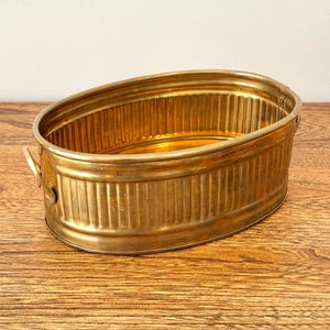 May include: An oval-shaped brass basin featuring a ribbed, vertical texture along its exterior walls. The container includes two small, curved handles attached to each side, with a polished metallic finish and a smooth, flat base.