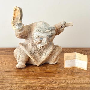 Peut inclure: Une sculpture en os sculpté, beige et poreuse, représentant une silhouette accroupie, les bras levés, tenant un objet plat dans une main et une petite tige dans l'autre. Une étiquette beige rectangulaire avec du texte est attachée.