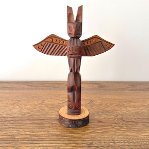 May include: A carved wooden totem pole with a bird-like figure at the top and outstretched wings. The dark brown sculpture is mounted on a small, round wooden base. The wings have detailed carved patterns.