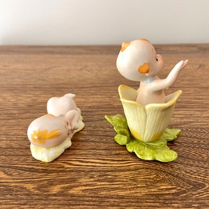 May include: Two porcelain figurines of babies, one sitting in a yellow flower and the other lying down on a yellow leaf. Both figurines have yellow hair and are wearing blue clothing.