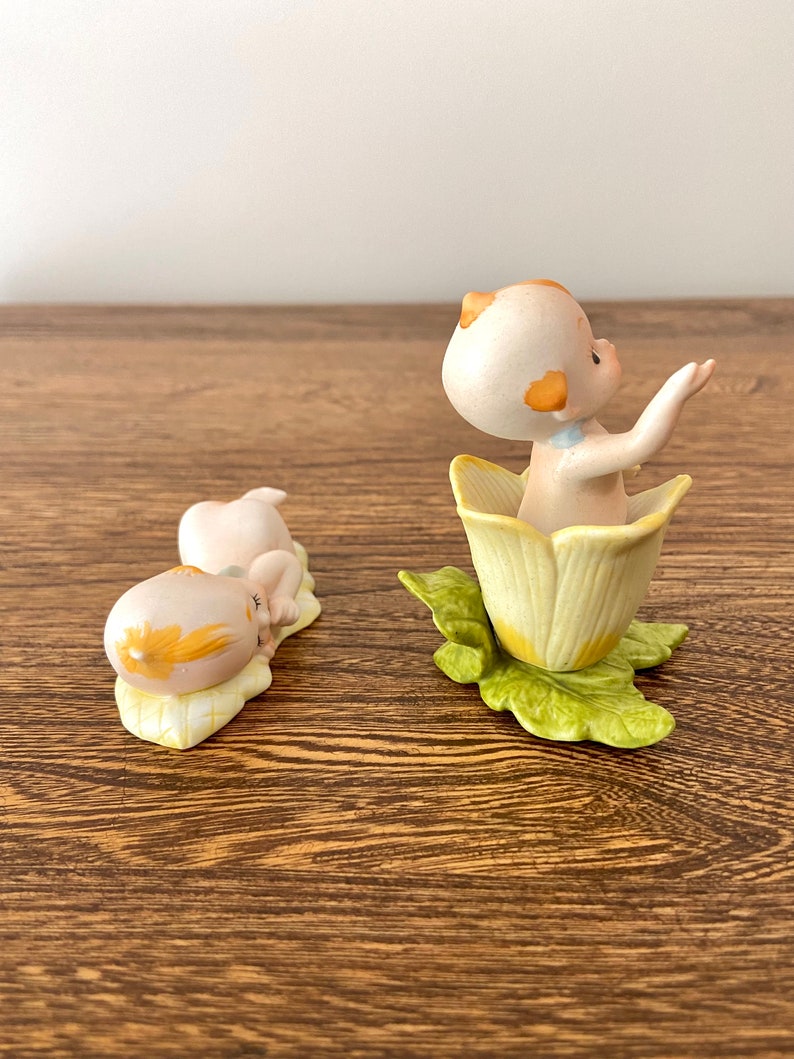 May include: Two porcelain figurines of babies, one sitting in a yellow flower and the other lying down on a yellow leaf. Both figurines have yellow hair and are wearing blue clothing.