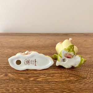 May include: Two porcelain figurines of a girl and a boy, both with blonde hair and wearing white dresses. The girl is holding a flower and the boy is holding a butterfly. The figurines are both sitting on a green leaf. The bottom of the figurines have the words "Little  Girls  Collection" and "Ganz  Designs" printed on them.