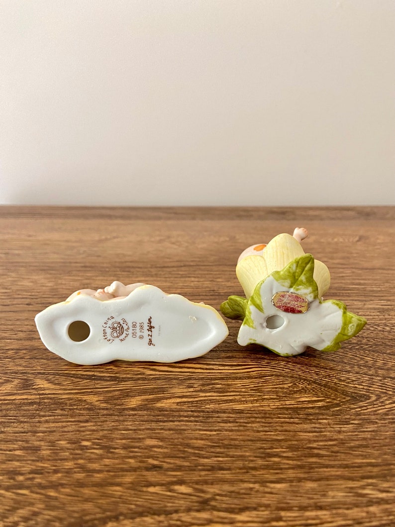 May include: Two porcelain figurines of a girl and a boy, both with blonde hair and wearing white dresses. The girl is holding a flower and the boy is holding a butterfly. The figurines are both sitting on a green leaf. The bottom of the figurines have the words "Little  Girls  Collection" and "Ganz  Designs" printed on them.