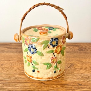 May include: A cylindrical ceramic biscuit barrel with a cream-coloured basketweave texture. The exterior features raised, hand-painted floral motifs in blue, orange, and pink with green leaves. It includes a matching lid and a curved wicker handle.