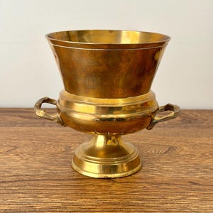 May include: A vintage brass urn with a flared top, two handles, and a pedestal base. The urn has a polished finish and is likely used for decorative purposes. The brass has a warm, golden hue, and the design is ornate.