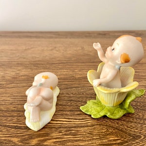May include: Two porcelain figurines of babies. One baby is lying on a yellow leaf, the other is sitting in a yellow flower.