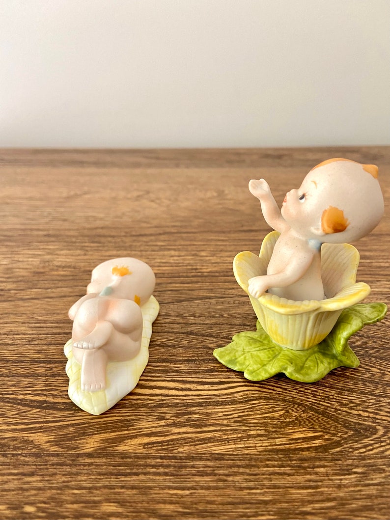 May include: Two porcelain figurines of babies. One baby is lying on a yellow leaf, the other is sitting in a yellow flower.