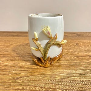 May include: White ceramic cup with a 3D floral design. The cup features a white flower with green leaves and a brown base.