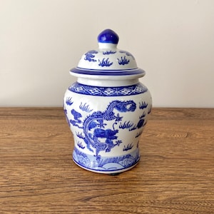 Puede incluir: Un jarrón de porcelana blanca con un diseño de dragón azul y blanco. El jarrón tiene una tapa y está decorado con nubes y un dragón.