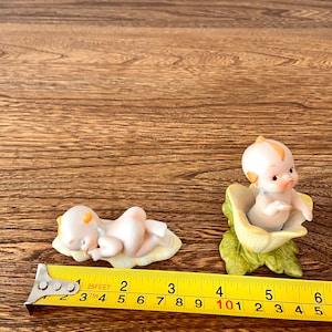 May include: Two porcelain figurines of babies, one lying down on a yellow and white blanket and the other sitting in a yellow flower. Both figurines have yellow hair and are wearing no clothes.