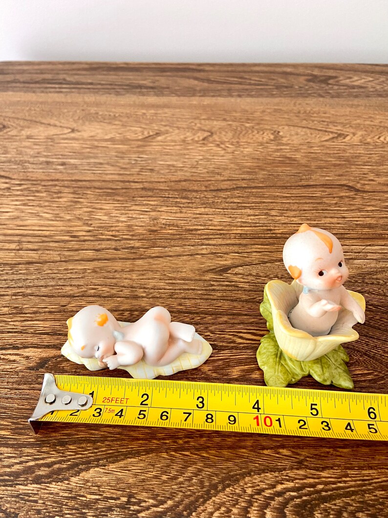 May include: Two porcelain figurines of babies, one lying down on a yellow and white blanket and the other sitting in a yellow flower. Both figurines have yellow hair and are wearing no clothes.