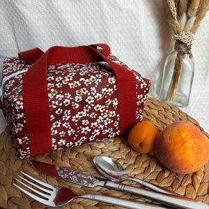 May include: A red lunch bag with a white floral pattern and red handles. The bag is sitting on a woven placemat with a fork, knife, spoon, and two peaches.