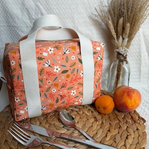 May include: A peach-colored lunch bag with a floral print. The bag has two white straps and a zipper closure. The bag is sitting on a woven placemat with a fork, knife, and spoon.