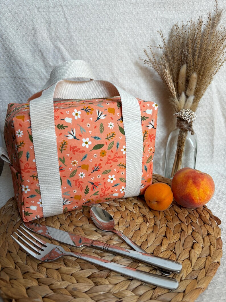 May include: A peach-colored lunch bag with a floral print. The bag has two white straps and a zipper closure. The bag is sitting on a woven placemat with a fork, knife, and spoon.
