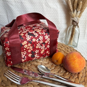 May include: A red lunch bag with a floral print and burgundy straps. The bag is sitting on a woven placemat with a fork, knife, spoon, and two peaches.